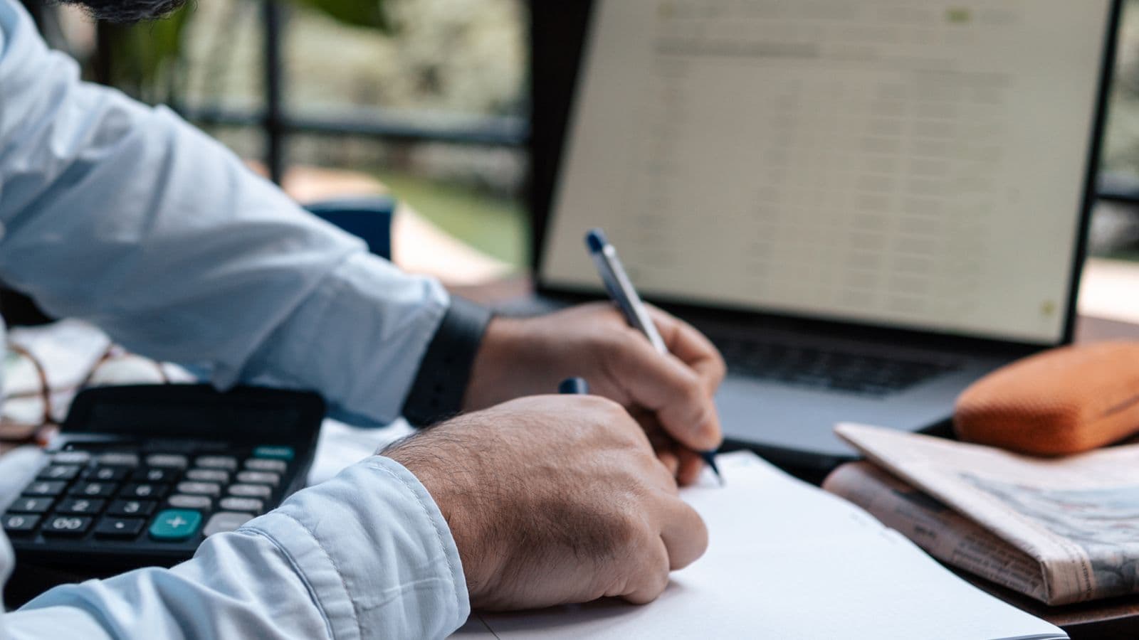 A person writing in a notebook beside a laptop and calculator at a desk, representing a fixed travel or work stipend